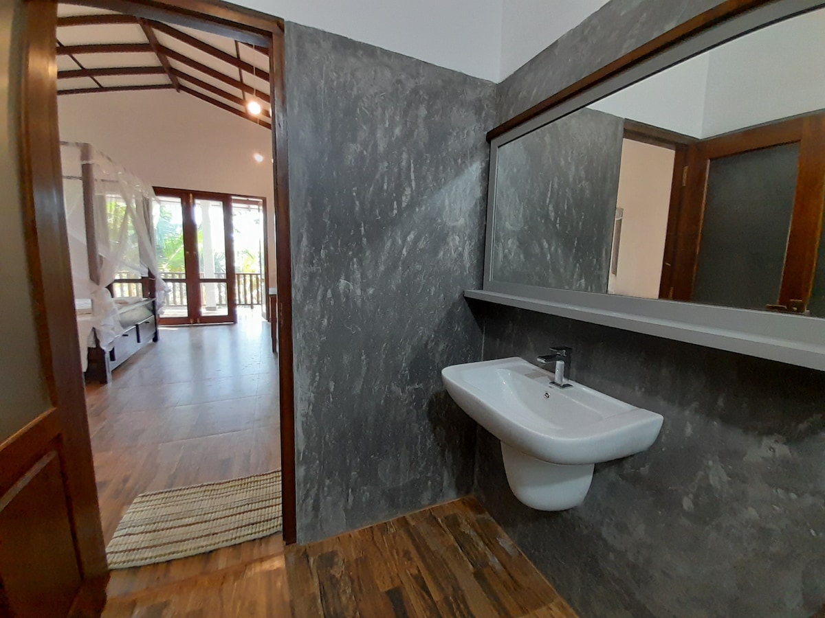 A spacious bathroom features a modern sink with a sleek faucet, set against a textured dark wall. Natural light streams in from a nearby doorway, illuminating the warm wooden floor. The transition to the adjoining bedroom area is visible through an open door.