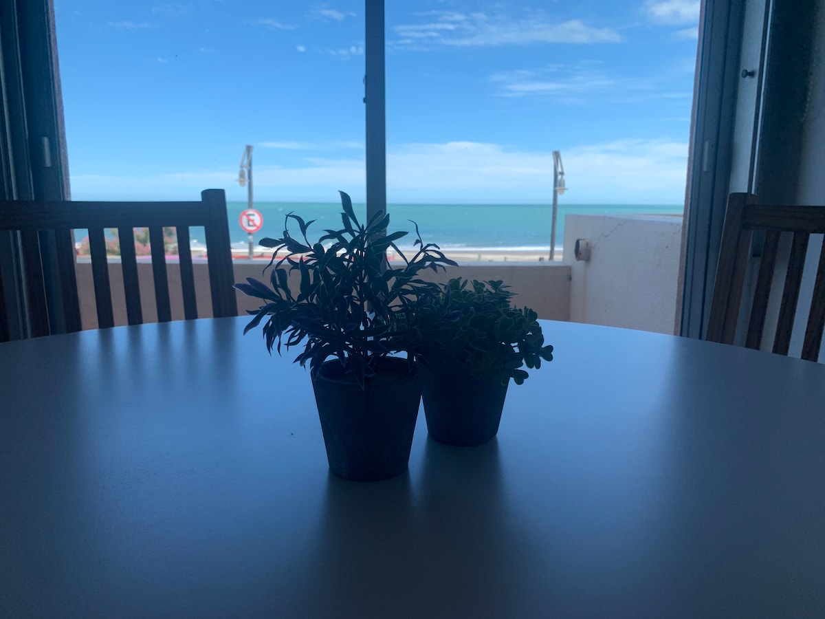 A round table is positioned near a large window, offering a view of the ocean and beach beyond. Two small potted plants create a focal point on the table, while natural light illuminates the space.