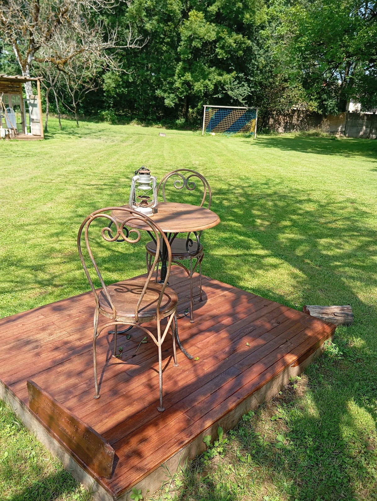 A wooden patio area is seen with a small round table and two metal chairs. The space is surrounded by lush green grass and trees, creating a serene outdoor setting. A decorative lantern rests on the table, providing a charming focal point.