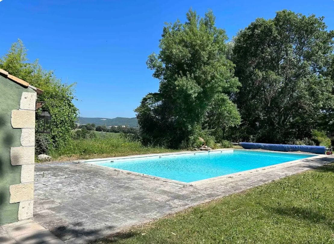An outdoor swimming pool is surrounded by a stone patio and greenery. Tall trees provide shade nearby, while a clear blue sky is visible above. The pool's azure water invites relaxation amidst the serene natural landscape.