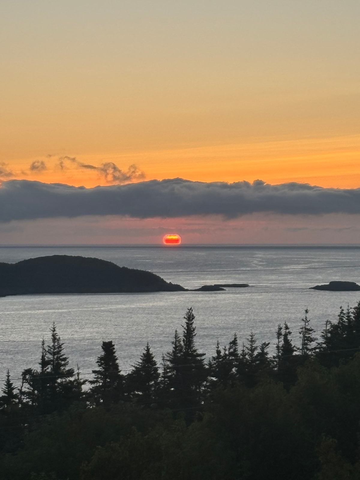 A sunset view is captured over a calm ocean, with the sun partially hidden behind clouds. Silhouettes of trees are visible in the foreground, leading down to rocky islands along the horizon, creating a serene and picturesque scene.