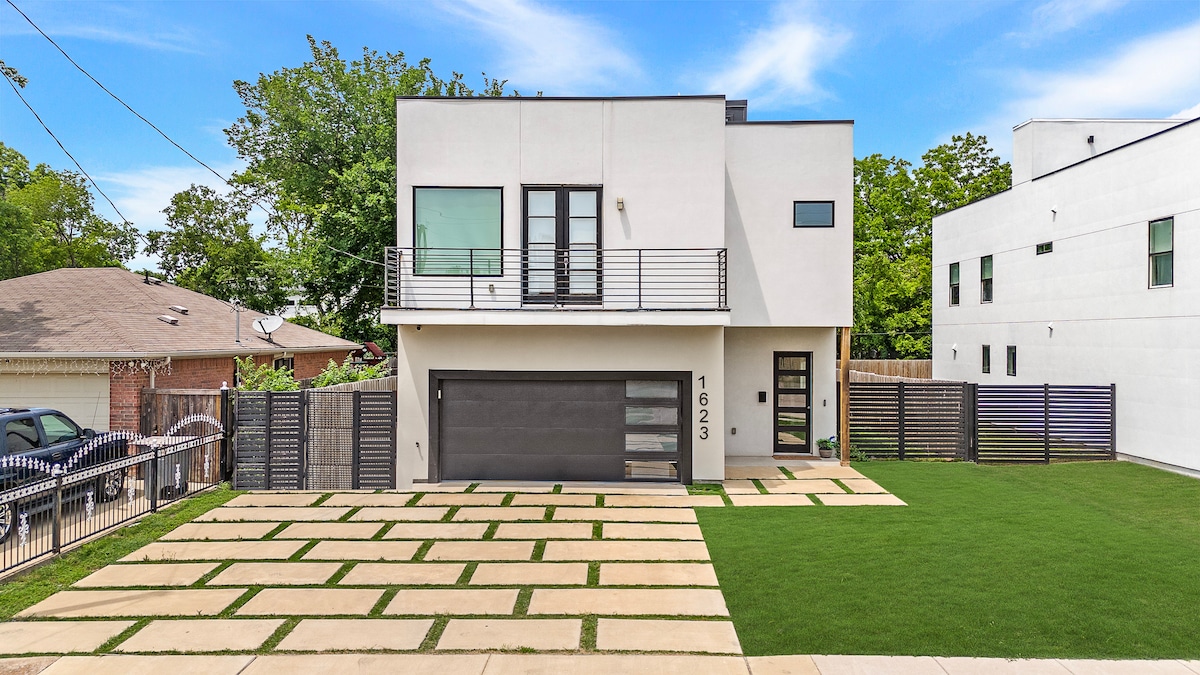 A modern two-story home is shown with a sleek facade featuring large windows and a contemporary design. The front yard is landscaped with green grass and stone paving, providing a welcoming appearance. A driveway leads to a spacious garage, offering convenient parking options.