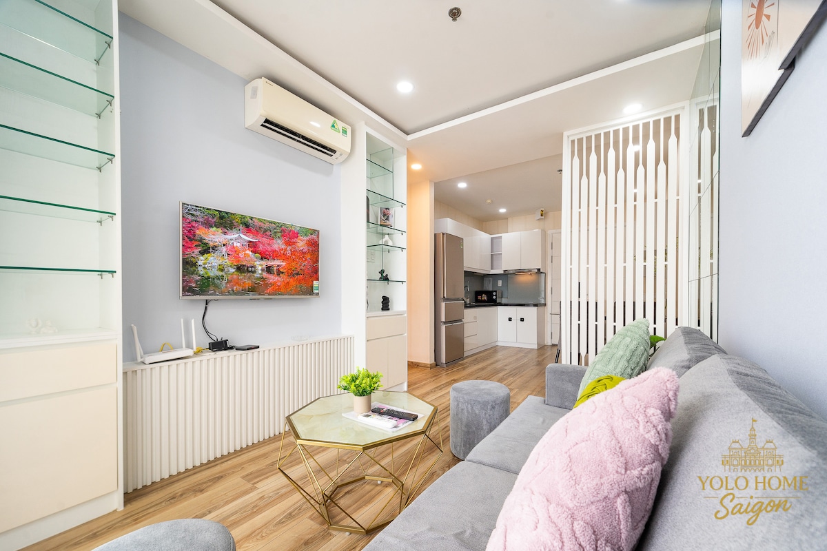 A modern living space features a comfortable gray sofa adorned with pillows, positioned next to a glass coffee table. A vibrant piece of wall art is displayed on the light-colored wall. The room is illuminated by natural light from large windows, enhancing the open feel.