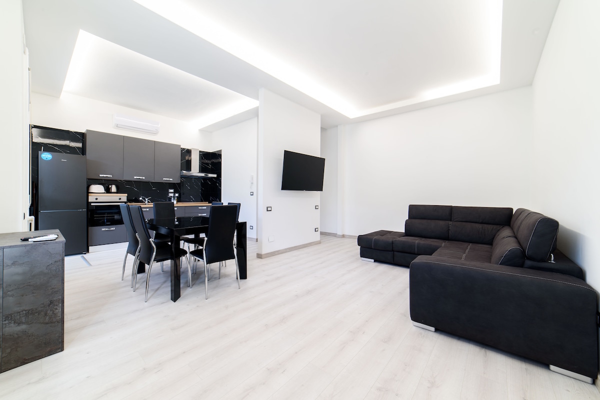 A spacious living area is visible, featuring a large sectional sofa in black and a dining table with black chairs. The modern kitchen is equipped with sleek black cabinetry and appliances. Natural light brightens the white walls, creating an open and airy environment.