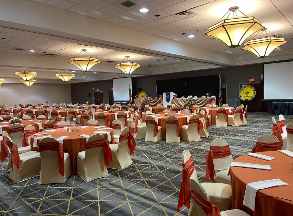 A spacious banquet room is arranged with round tables covered in orange linens, complemented by light-colored chair covers with orange sashes. Multiple chandeliers illuminate the area, with presentation screens positioned at the front, creating an inviting atmosphere for events.