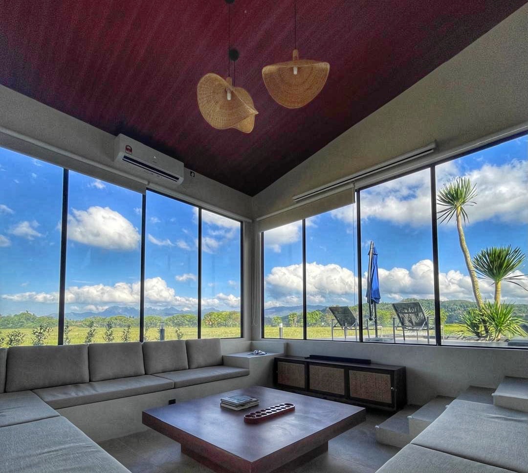 A spacious living area features large windows that frame views of the surrounding greenery and distant mountains. Natural light fills the room, highlighting a sectional sofa and a modern coffee table at the center. Decorative pendant lights hang from the high ceiling.