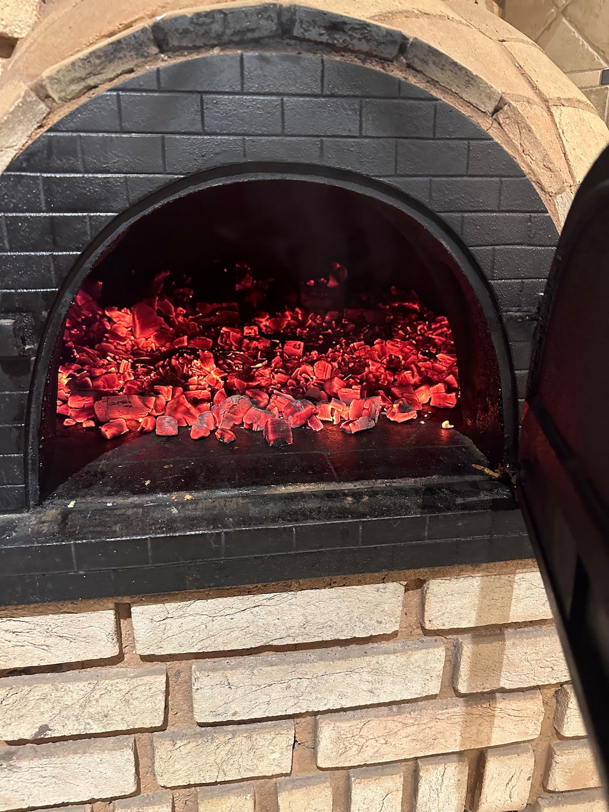 A traditional outdoor cooking oven is visible, featuring glowing red coals and a brick facade. The archway opening reveals the heat radiating from within, indicating readiness for cooking. The textured stone surrounding the oven adds to the rustic ambiance.