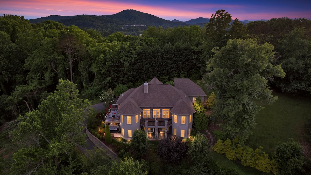 The exterior of the luxurious mansion is showcased, surrounded by lush greenery and trees. The setting sun casts a warm glow on the home, highlighting large windows that reflect the natural beauty of the mountains in the background.