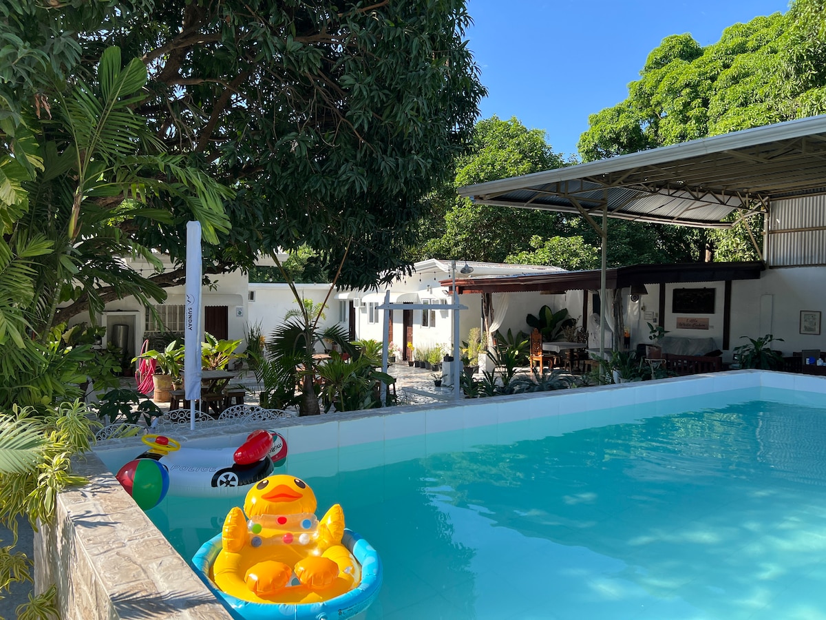 A tranquil outdoor area features a swimming pool with a bright yellow inflatable duck floating on the water. Lush greenery surrounds the pool, providing shade. In the background, relaxation spaces are visible under a covered terrace, showcasing a welcoming environment for guests.