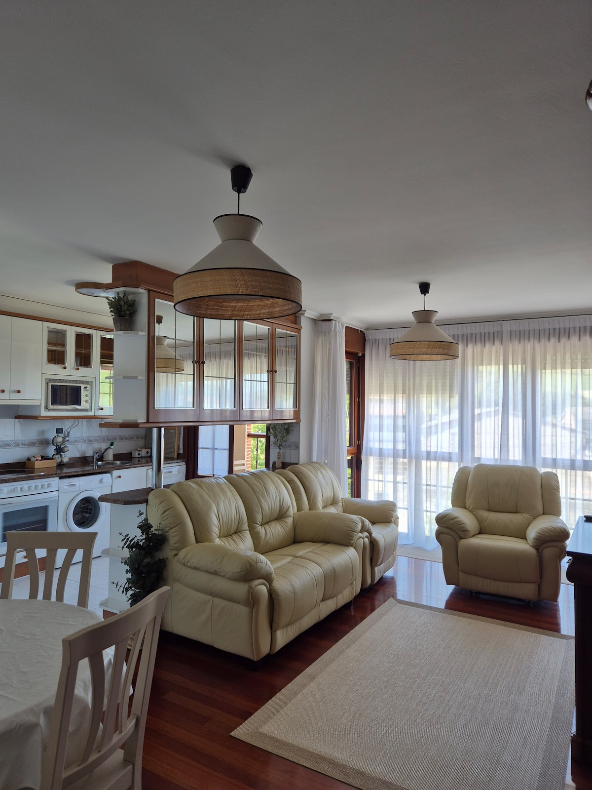 The living area features a comfortable cream-colored sofa and armchair, set against a backdrop of large windows allowing natural light. A dining table with white chairs occupies one corner, while soft lighting is provided by two woven pendant lamps hanging from the ceiling.