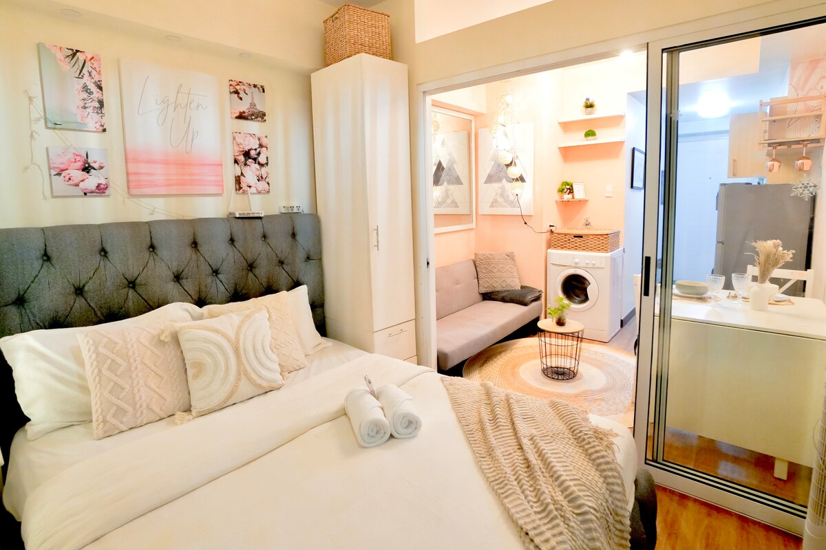 A cozy sleeping area features a tufted gray headboard against neutral-colored walls. A queen bed is neatly dressed with white linens and decorative pillows. In the background, a washing machine is visible, alongside a compact seating area with a gray sofa, creating an inviting ambiance.