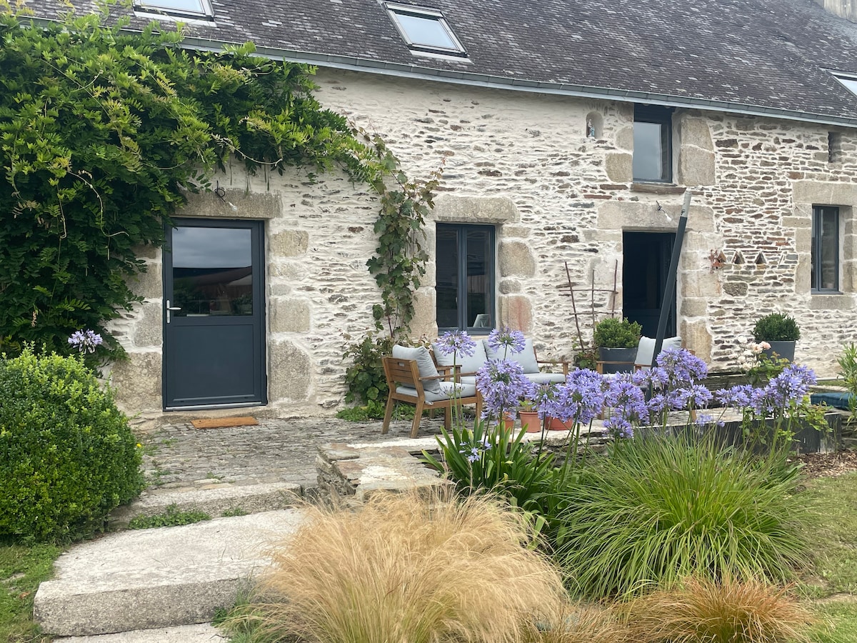 The exterior of a stone house features a welcoming entrance with a modern gray door. Lush greenery and vibrant purple flowers surround a cozy seating area, composed of wooden chairs and a small table. Stone pathways lead through the garden, enhancing the serene atmosphere.