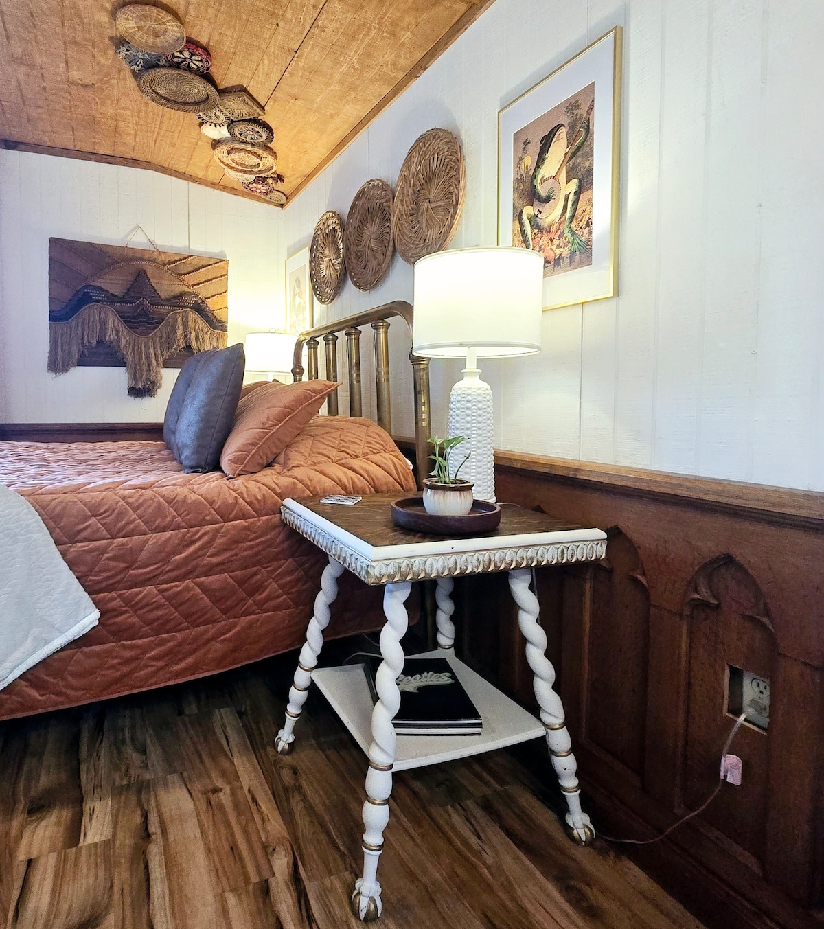A cozy corner of a bedroom features a wooden bed with warm bedding and decorative pillows. A white lamp illuminates a small, intricately designed side table, which holds books. The wall is adorned with woven art, adding character to the inviting space.