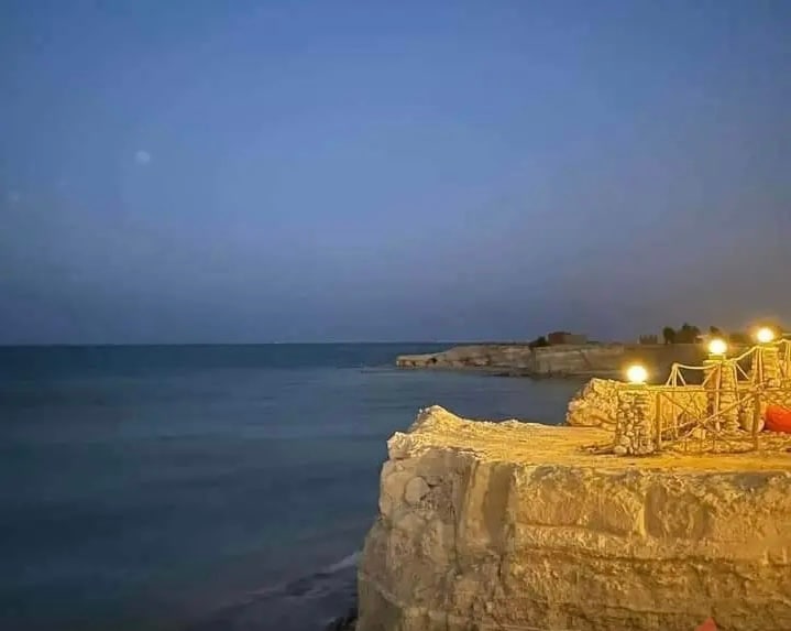 A rocky shoreline is visible under the evening sky, where gentle waves meet the coast. Soft lighting from nearby outdoor seating casts a warm glow, enhancing the peaceful atmosphere of the coastal setting.