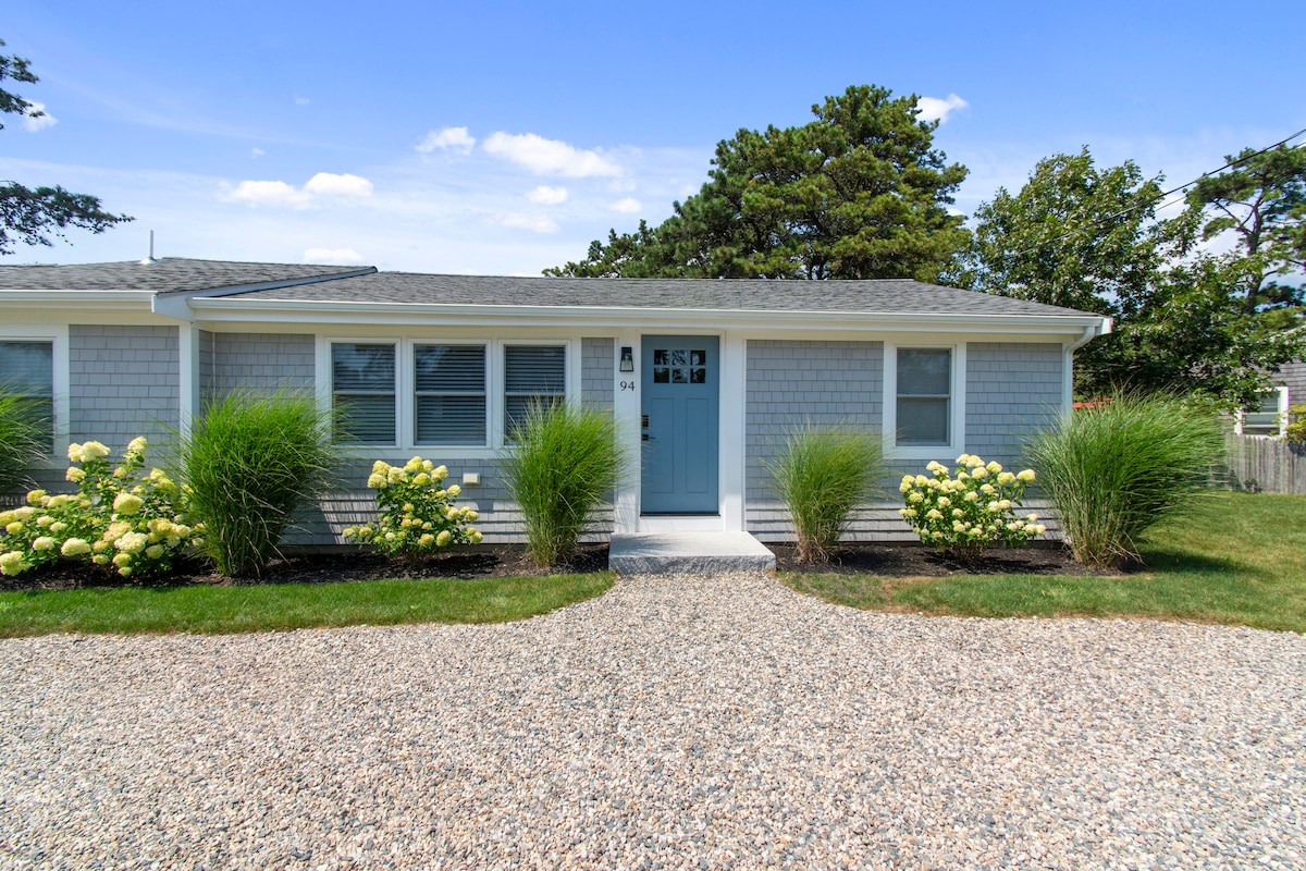The entrance to a renovated duplex cottage is visible, featuring a light blue front door framed by lush greenery and flowering bushes. A gravel pathway leads to the door, with tidy landscaping adding to the welcoming appearance of the property.