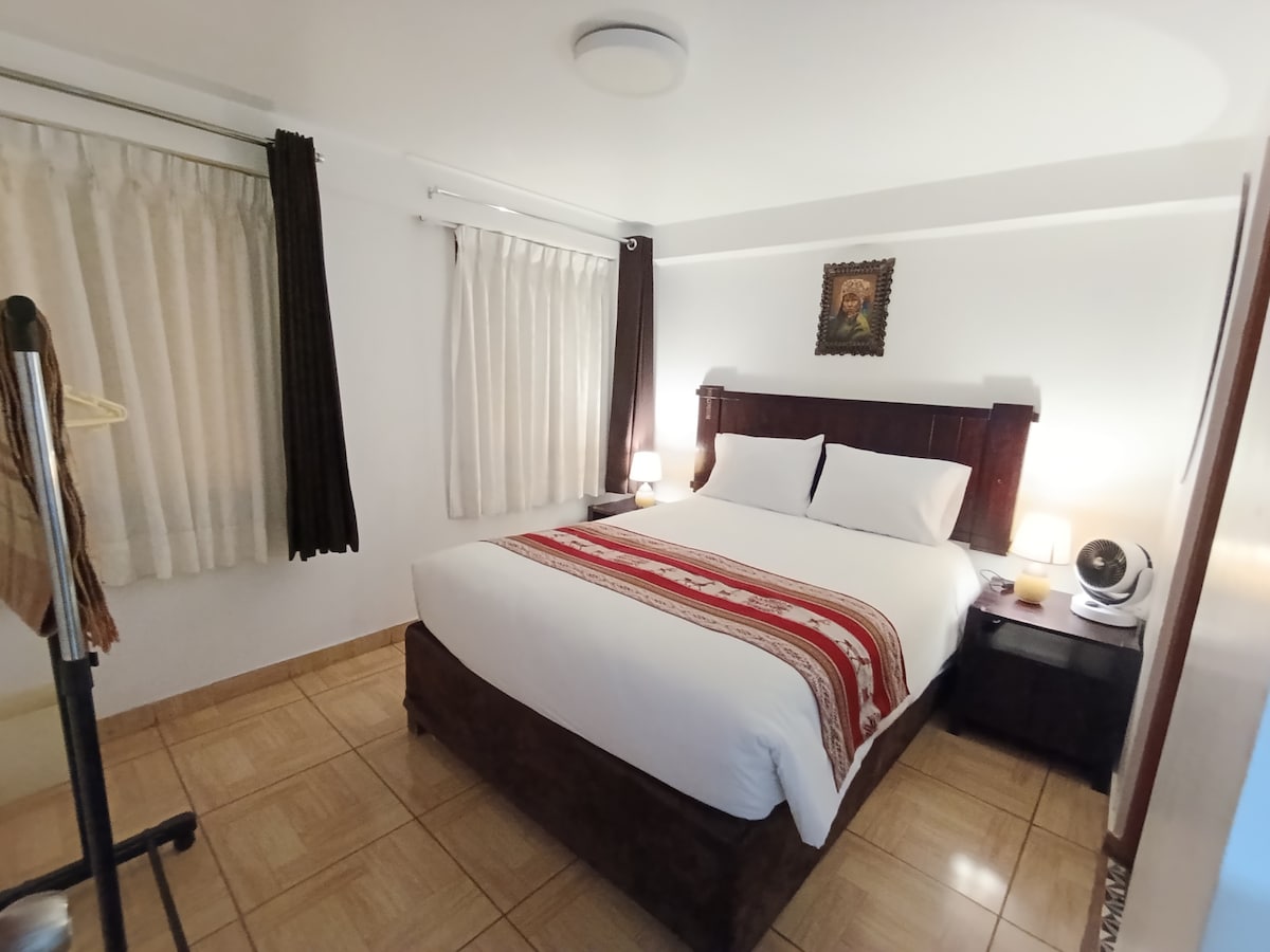 A well-lit bedroom features a spacious bed dressed in white linens, adorned with a decorative striped throw. Two bedside tables with lamps flank the bed, while sheer and dark curtains frame the windows. A simple coat rack is positioned near the wall.