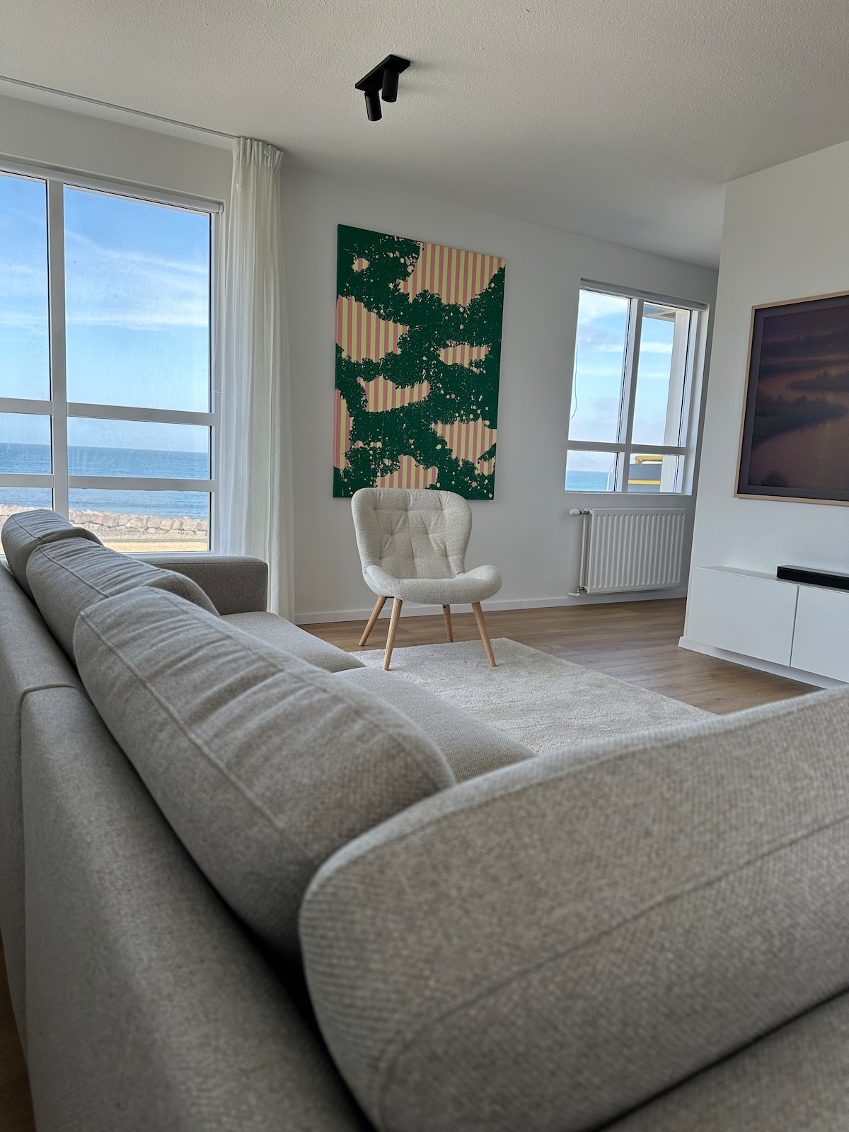 A modern living area is showcased, featuring a light grey sofa positioned towards large windows that offer a view of the sea. A green and beige artwork adorns the wall, while a single white chair complements the space. Natural light fills the room.