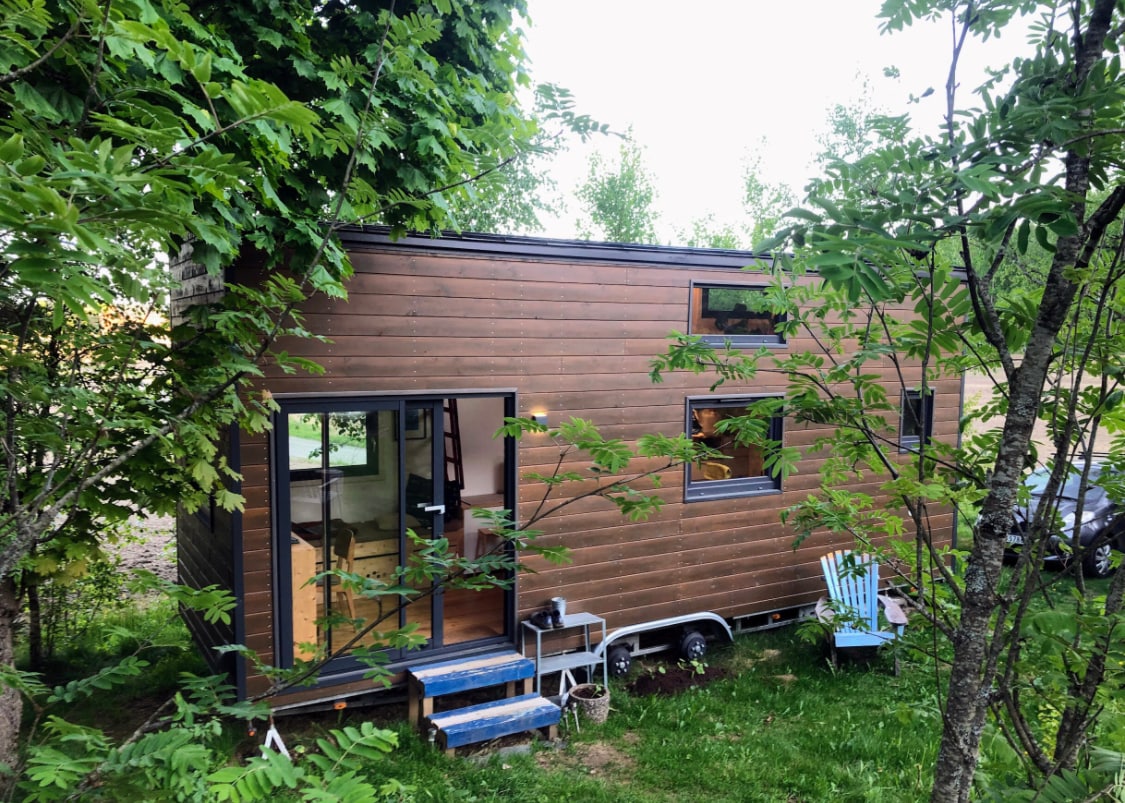The tiny house is nestled among trees, featuring a wooden exterior with several windows that provide natural light. Blue steps lead to an entrance, while a few outdoor chairs are visible nearby, set against a lush green backdrop.