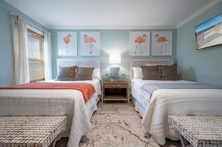 Cozy twin bedroom with flamingo art, soft lighting, beach decor, and charming vibes.