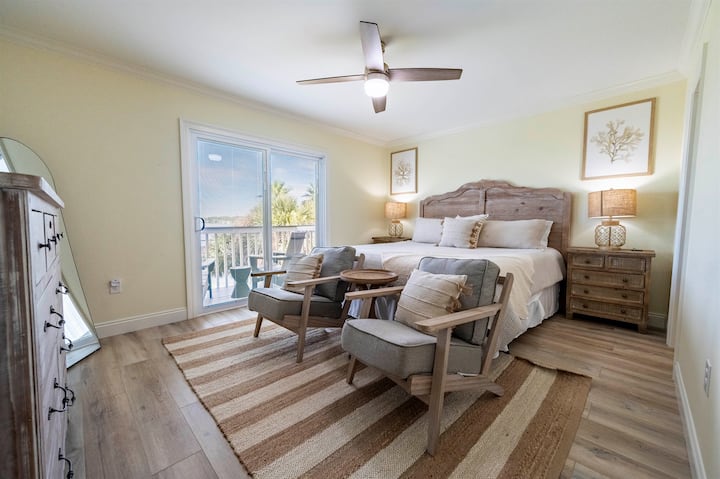 Spacious upstairs master suite with a king bed, warm wood furnishings, and soft neutral tones. Opens to a balcony overlooking Little Lagoon, with seating for quiet moments and fresh coastal air.