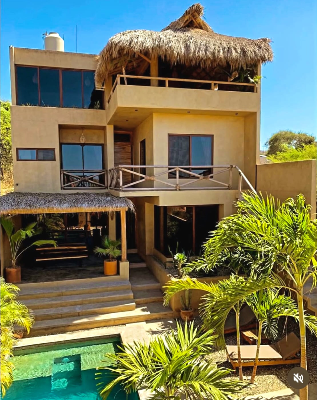 The house is showcased from an outdoor perspective, highlighting its three levels with a thatched roof and large windows. Lush greenery surrounds the property, framing a private pool below. A covered patio area with ample seating is visible, inviting relaxation in a serene environment.