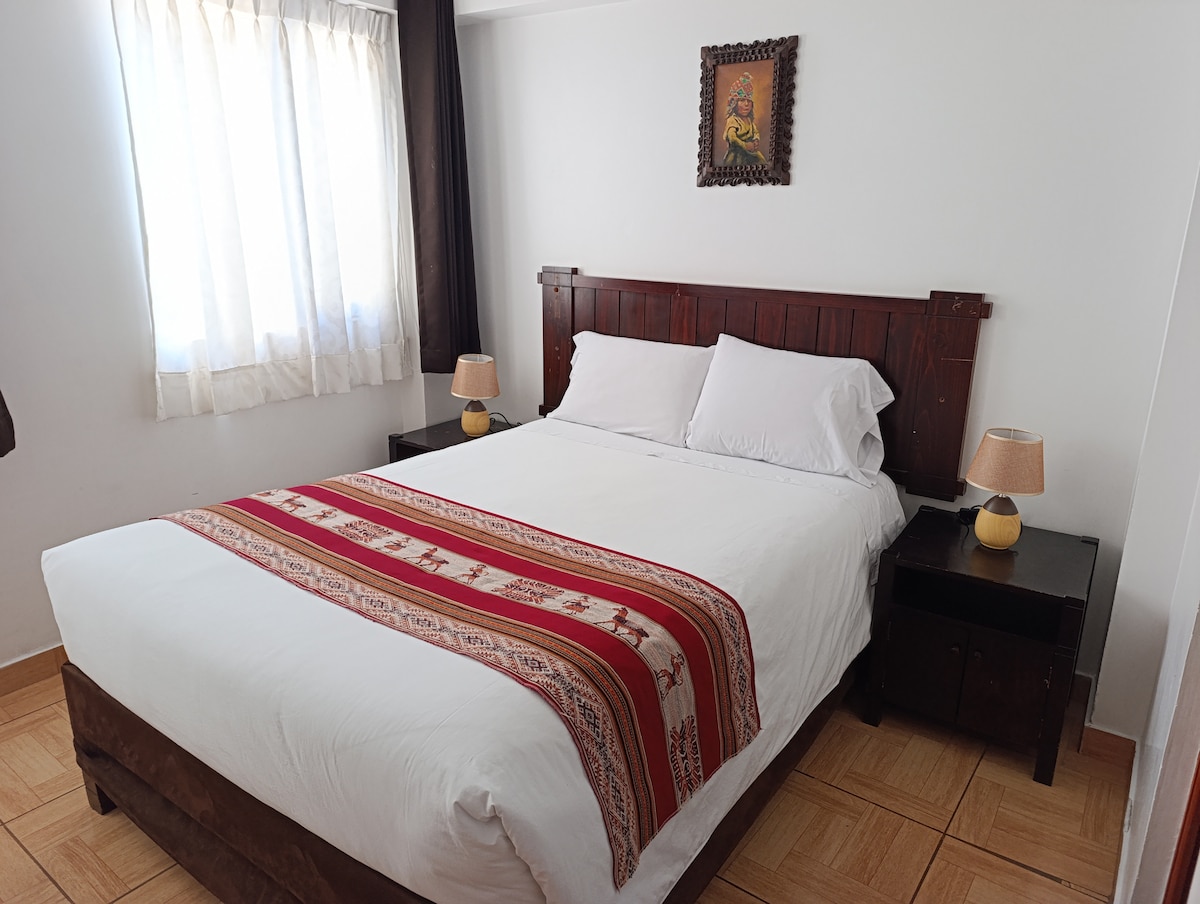 A bright and airy bedroom features a spacious bed with a white duvet cover and decorative throw. Dark wooden nightstands with lamps are positioned on either side. Natural light filters through sheer curtains, illuminating the room's neutral color palette.