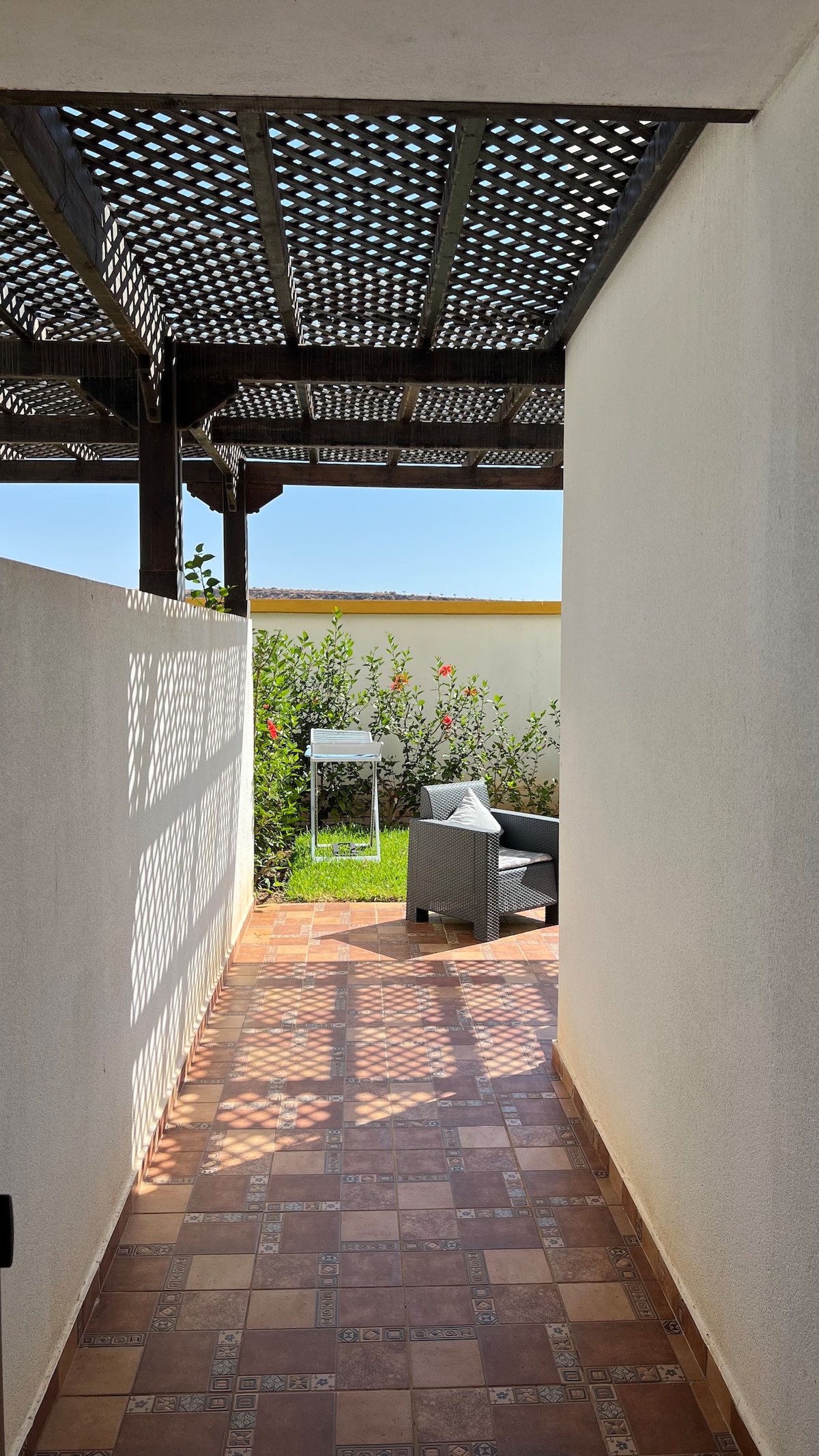 A corridor leads to a serene outdoor space, featuring sunlight filtering through a trellis overhead. A comfortable seating area is visible, surrounded by greenery and flowers, creating a private retreat.