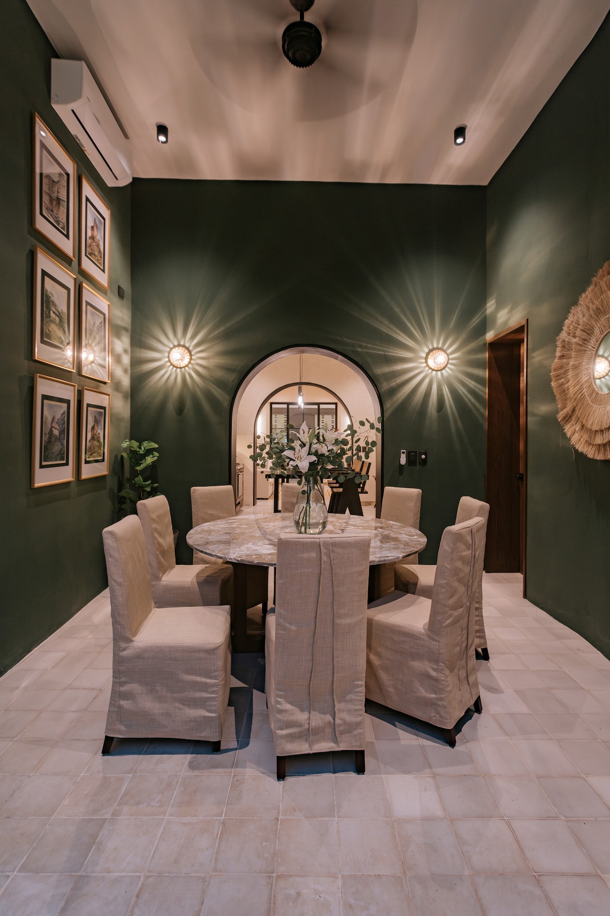 A dining area features a round table surrounded by six upholstered chairs. The walls are painted a deep green, complemented by framed artwork. Soft lighting highlights the space, while a small potted plant adds a touch of greenery.