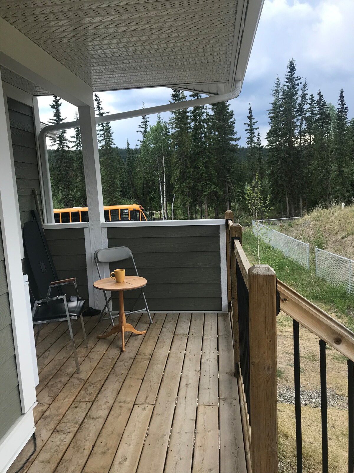 A wooden deck is depicted with a small round table and a single chair positioned on it. A yellow bus is visible in the background, surrounded by lush trees. The area is characterized by a peaceful setting, with grass and a fence surrounding the property.
