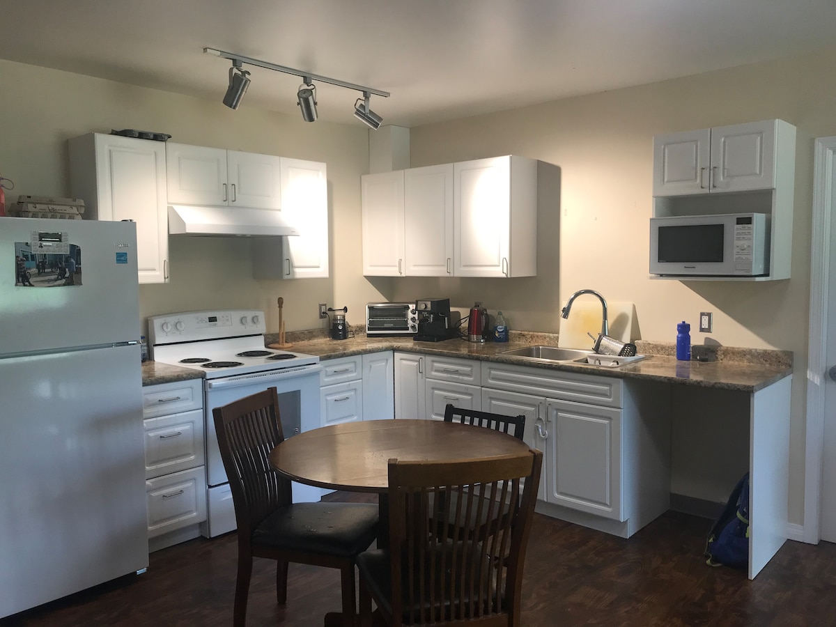 A bright kitchen area is equipped with white cabinetry and granite countertops. A round wooden table with four chairs is positioned in the center. Major appliances, including a refrigerator, stove, and microwave, are neatly arranged, contributing to a functional culinary space.