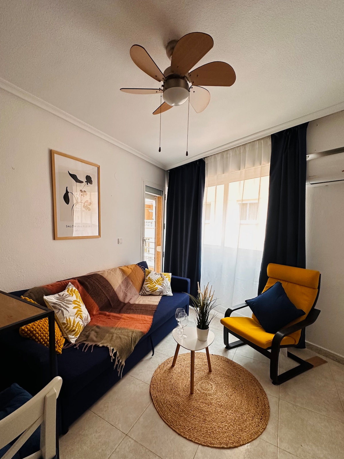 A cozy living area features a navy blue sofa adorned with bright throw pillows and a colorful blanket. A round table holds two glasses and a small plant. A yellow armchair complements the space, while sliding glass doors introduce natural light, framed by dark curtains.