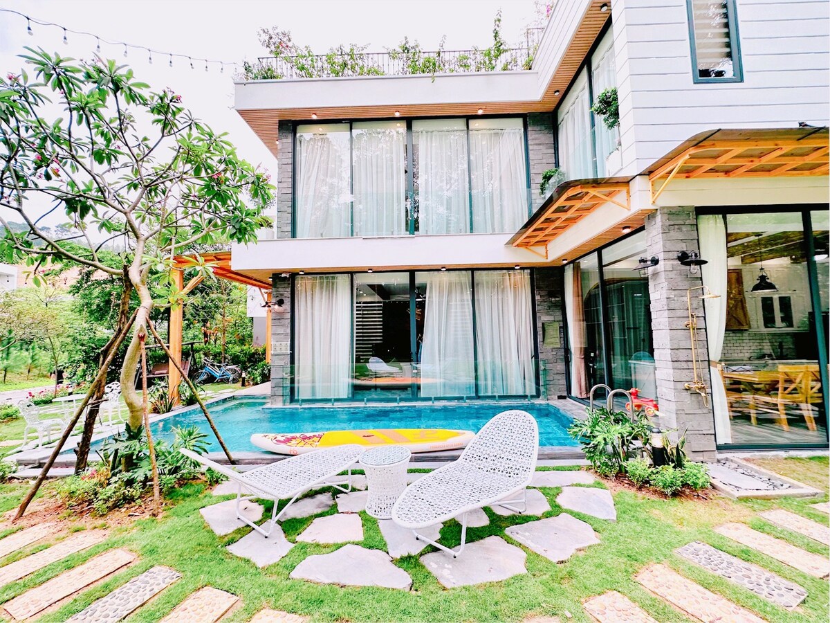 A contemporary villa exterior is surrounded by lush greenery, featuring a tranquil blue pool. Comfortable lounge chairs are positioned on a stone pathway, inviting relaxation. Large windows reveal a bright interior, seamlessly integrating outdoor and indoor spaces.
