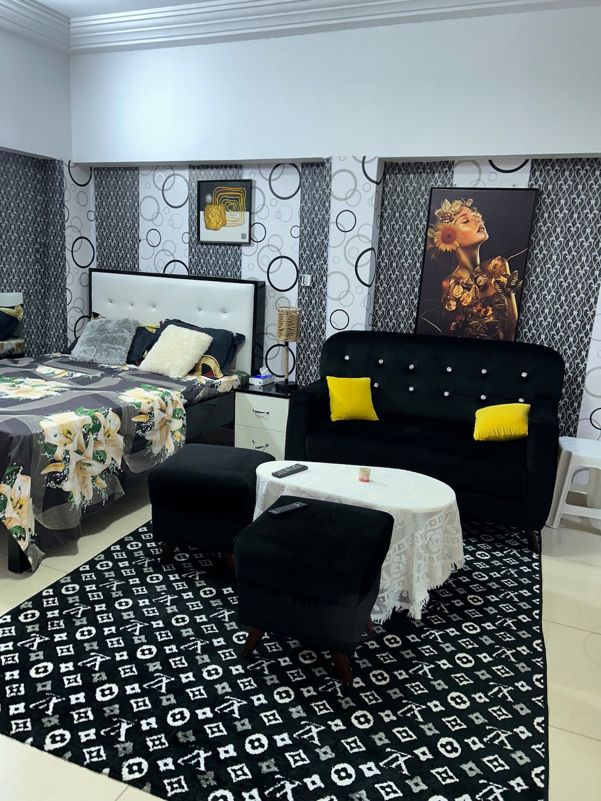 A modern bedroom is showcased with a spacious bed adorned in a floral-patterned blanket. A cozy black sofa with decorative yellow cushions faces a small coffee table, complemented by a geometric patterned rug. Stylish wallpaper features circular designs, adding to the contemporary decor.