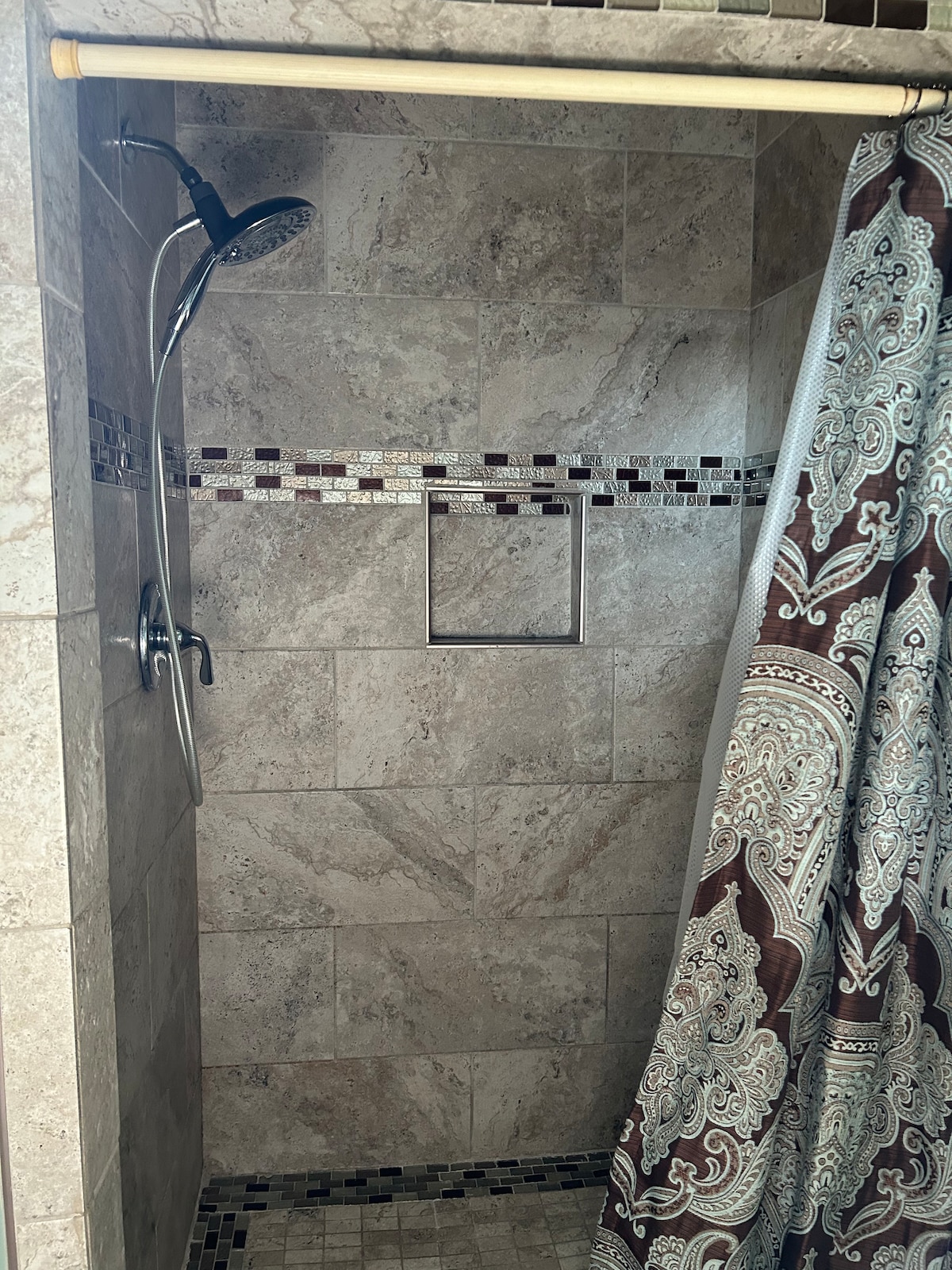 A spacious shower area is defined by neutral stone tiles. A handheld showerhead is mounted on the wall, accompanied by a decorative shower curtain featuring an intricate pattern. A built-in niche for toiletries is located on one side.