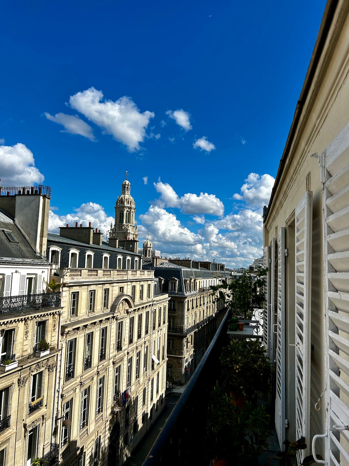 A view from the balcony showcases a bustling Parisian street lined with elegant buildings. A tall historic clock tower rises in the distance against a backdrop of vibrant blue sky and fluffy white clouds.
