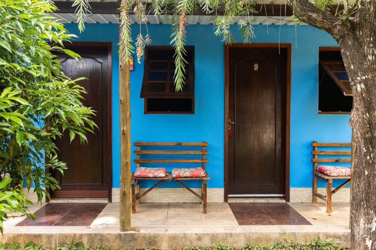 Two doorways, marked 9 and 11, are framed by vibrant blue walls in a garden setting. Wooden benches with floral cushions are positioned on a tiled patio, complemented by lush green foliage and a tree nearby.