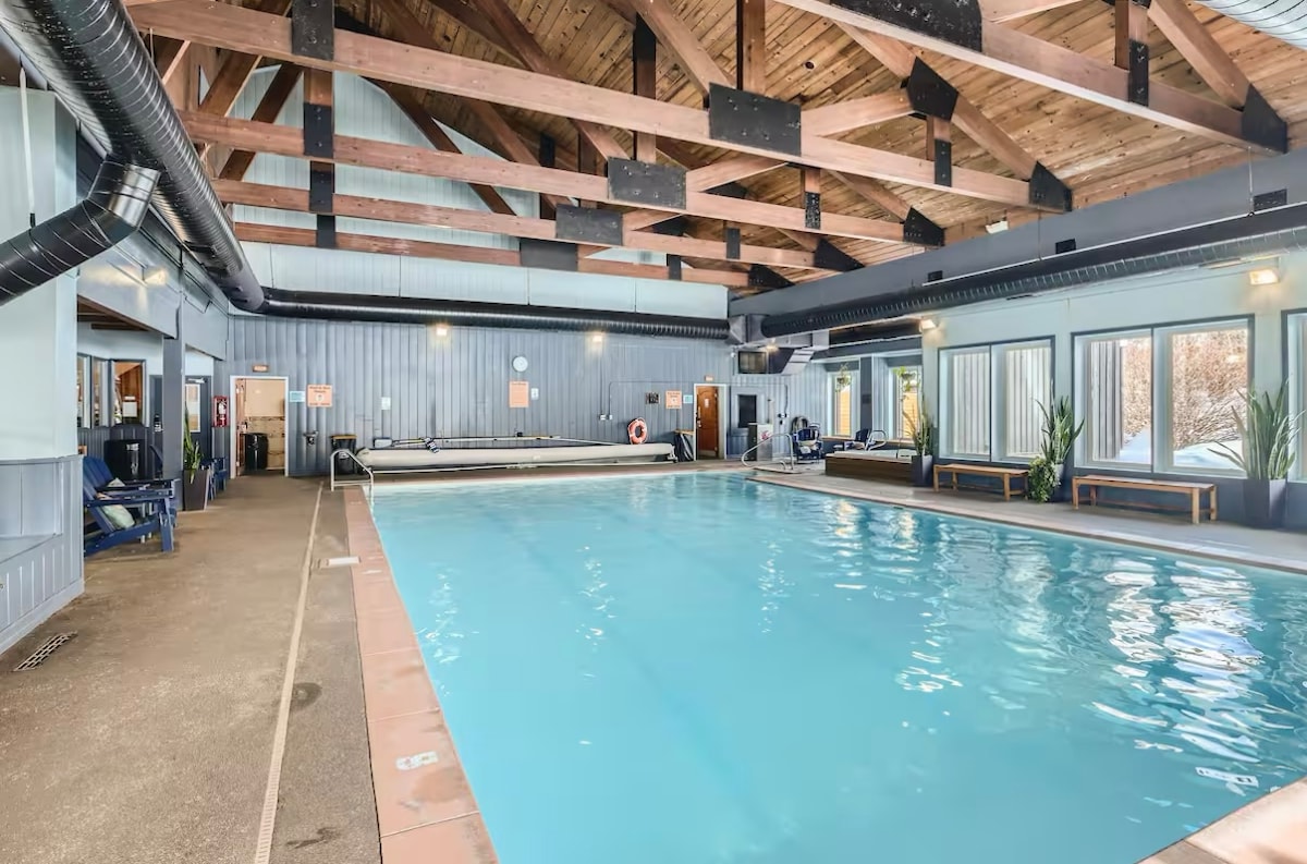 An indoor pool area is shown with a clear, blue swimming pool beneath a large wooden trussed ceiling. Natural light is pouring in through several windows, while comfortable seating is placed along the walls. Lifeguard equipment can be seen for safety.