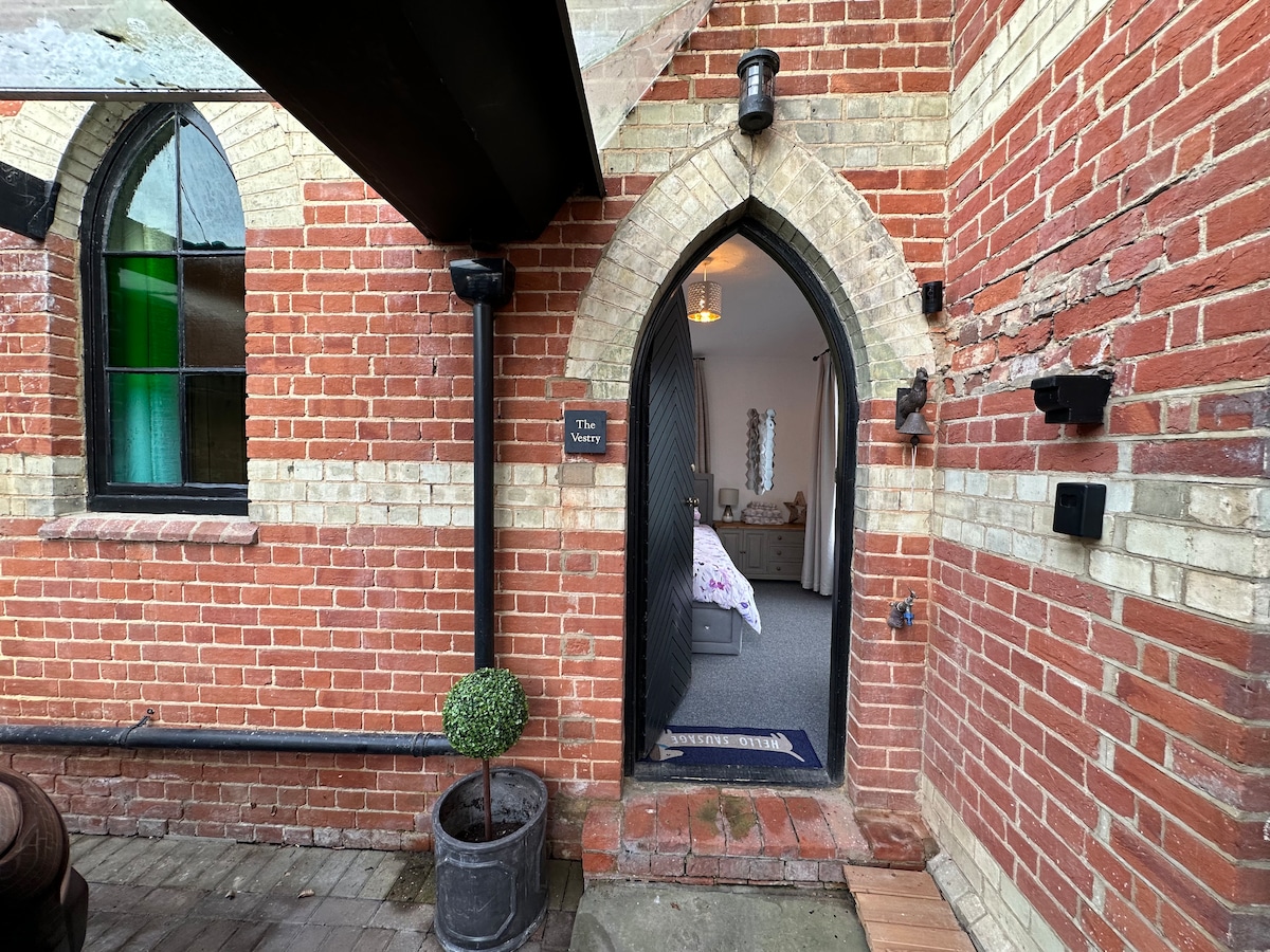 An arched entrance framed by red brick leads to a cozy interior. A potted plant sits beside the doorway, while the interior space is visible, showcasing soft furnishings and gentle lighting within.