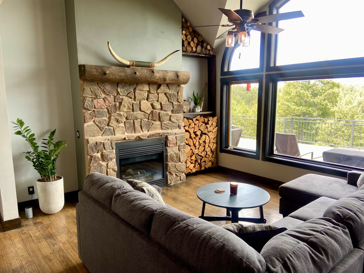 A warm and inviting living area features a stone fireplace with a wooden mantle. Large windows provide natural light, offering views of the surrounding greenery. A cozy sectional sofa surrounds a small coffee table, while a decorative plant adds a touch of nature to the space.