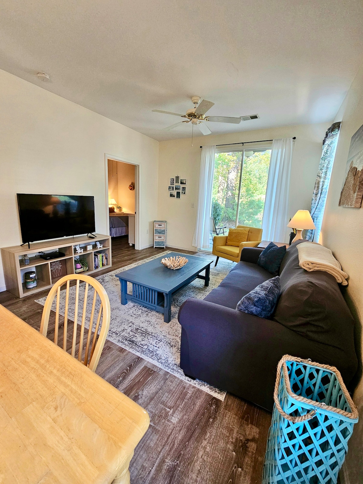 A welcoming living area features a dark sofa with patterned cushions and a coffee table at its center. A TV is situated on a light wood console. Natural light fills the space through large windows, accompanied by light curtains. Adjacent, a dining table with wooden chairs offers additional seating.