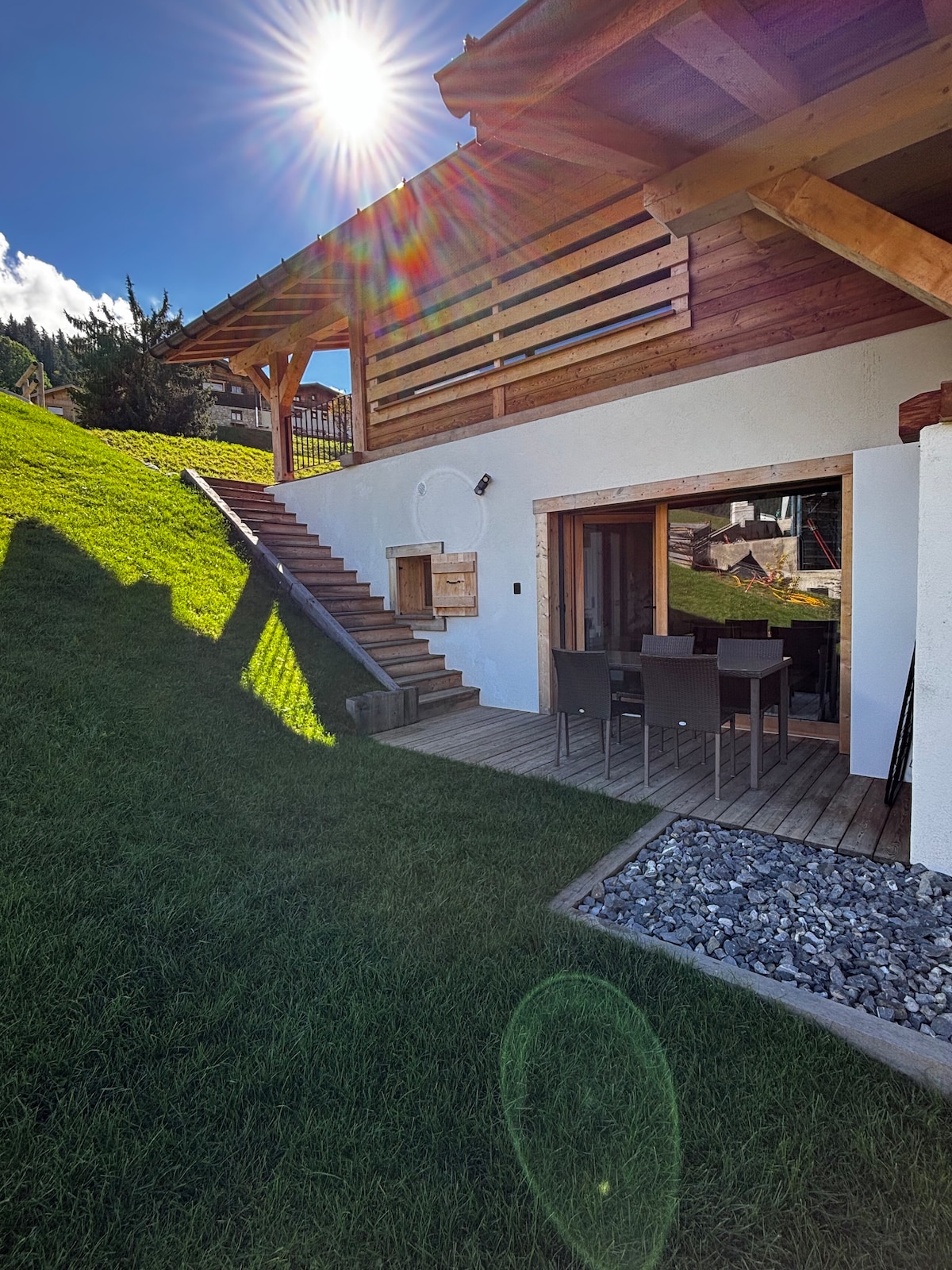 An exterior view of the chalet highlights a grassy slope leading to a wooden terrace. The terrace features a dining table with chairs, and large glass doors open into the living space. Sunlight creates a bright atmosphere, enhancing the natural surroundings.
