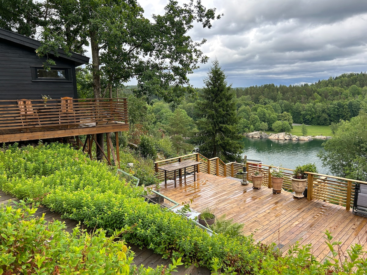 A spacious wooden terrace is complemented by a dining table and several planters, surrounded by lush greenery. The serene water and trees in the background create a tranquil setting. A dark wooden structure with a deck is visible above, enhancing the natural atmosphere.