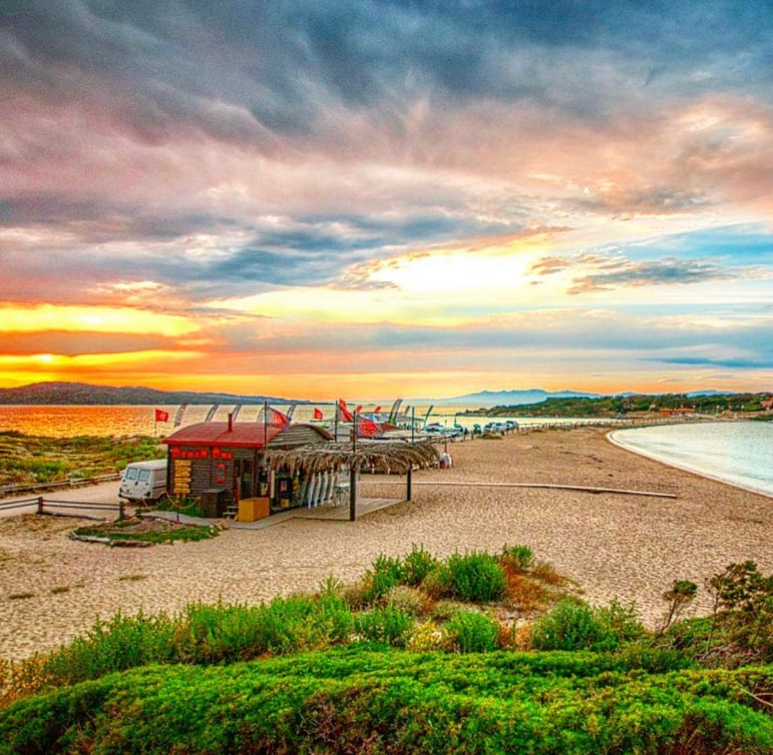 A sandy beach stretches towards the horizon, framed by gentle waves and greenery. Several beach huts line the shore, offering shaded spots under thatched roofs. A colorful sunset casts warm tones across the sky, reflecting on the water's surface in the distance.