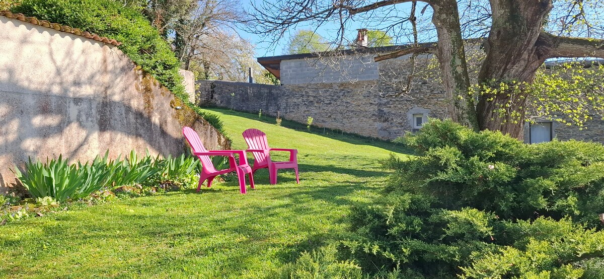 A serene garden area is depicted, featuring two vibrant pink chairs positioned on a lush green lawn. Surrounding flowers and foliage add to the natural setting, while a stone wall and trees provide a sense of privacy and tranquility.
