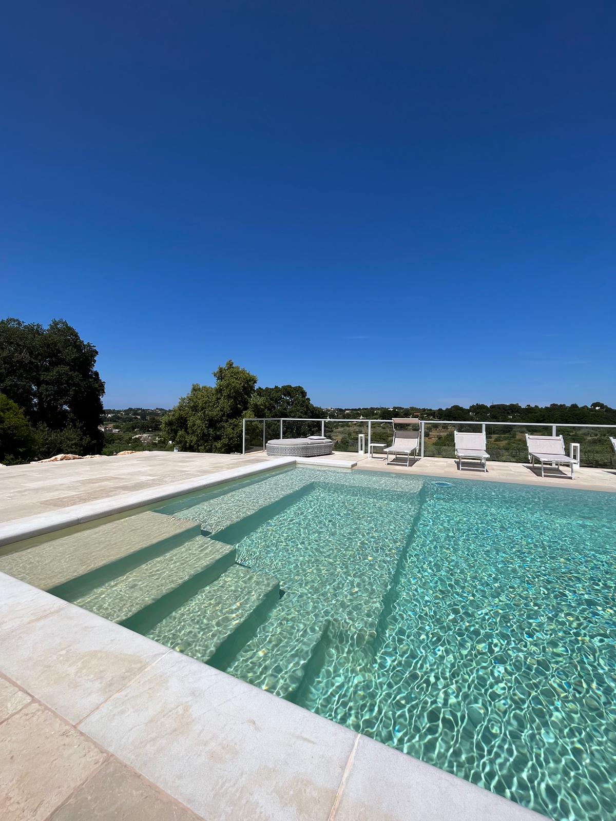 A serene swimming pool is depicted, featuring clear turquoise water and shallow steps leading in. Surrounding chairs are positioned on the pool deck, offering a space for relaxation. Lush greenery and a clear blue sky extend in the background, contributing to a tranquil outdoor setting.