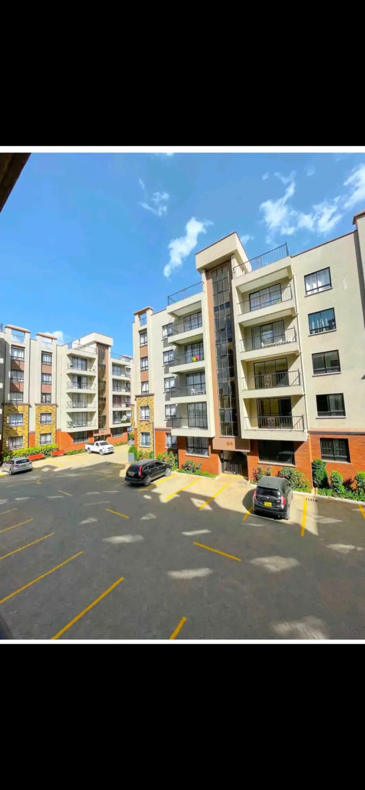 A modern apartment building showcases a clean architectural design with multiple stories and large windows. A parking lot in the foreground features several cars, while landscaped greenery accentuates the property. The clear blue sky forms a bright backdrop for the scene.