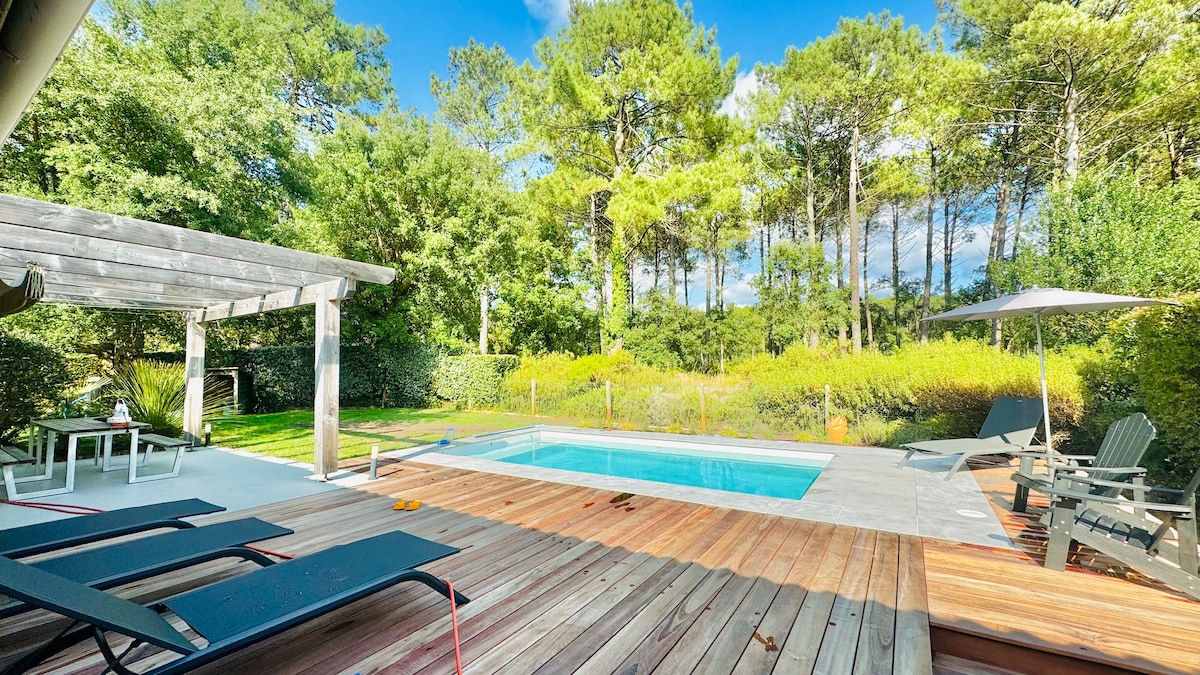A serene outdoor area is presented, featuring a heated swimming pool surrounded by a concrete deck. The space includes a wooden terrace with lounge chairs and a shaded seating area. Lush greenery and tall trees frame the backdrop, contributing to a tranquil atmosphere.