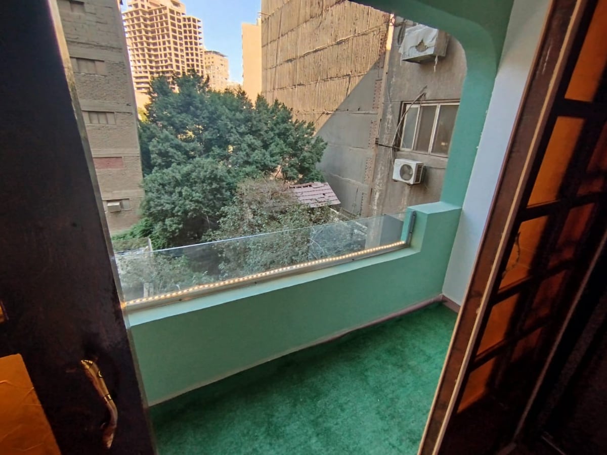 A balcony is shown with a green carpeted floor and a glass railing providing an unobstructed view of surrounding trees and nearby buildings. Natural light fills the space, creating a bright atmosphere.