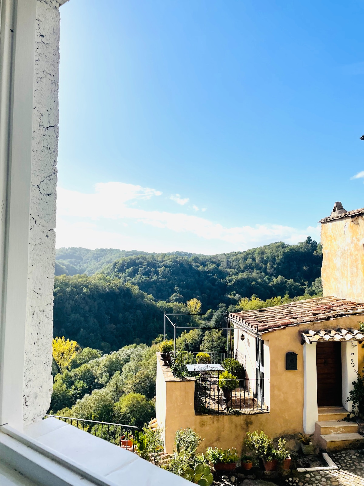 A scenic view is captured through a window, revealing lush green hills and a clear blue sky. The landscape features a mix of trees and plants, while the neighboring building showcases a terracotta roof and potted plants on the balcony.