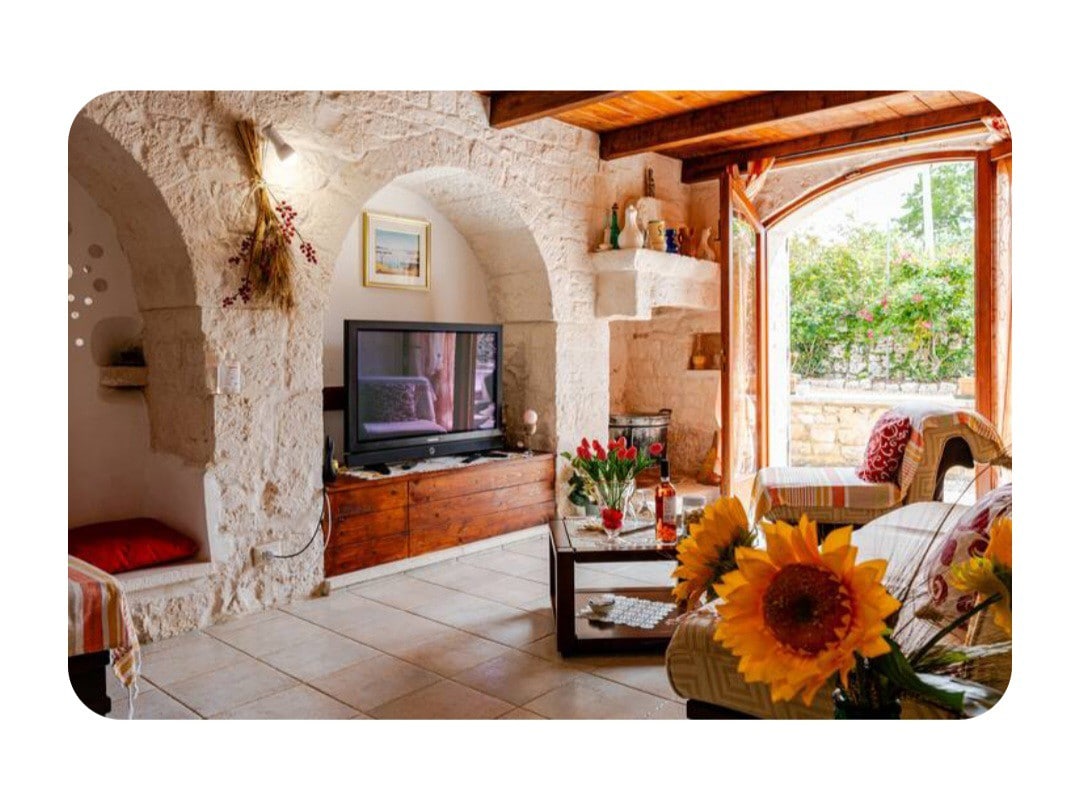 A cozy living area features a large television mounted on a wooden console, with a stone wall creating a rustic ambiance. Natural light floods through an arched window, illuminating comfortable seating adorned with floral decor. A small seating space is visible to the left.